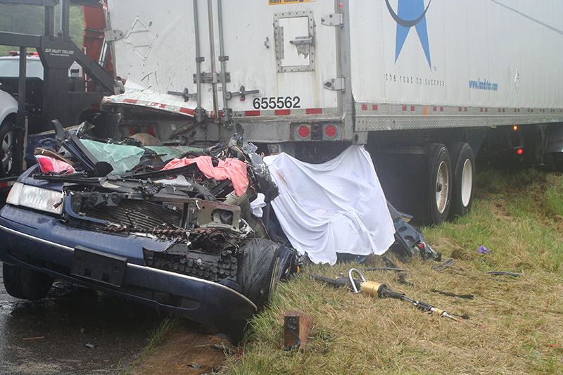 Photo of our blue Ford Crown Victoria car, with the rear of the car forced under the back of the truck trailer from the collision. Most of the car is nearly unrecognizable from the damage There is a sheet covering part of the car. There is blood on the sheet.